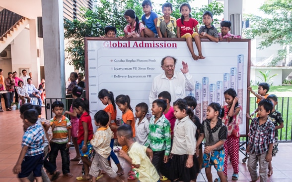Beat Richner in Cambogia nel 2013, circondato da bambini.Dietro di lui, una lavagna che illustra il numero di bambini curati negli ospedali, anno dopo anno. 