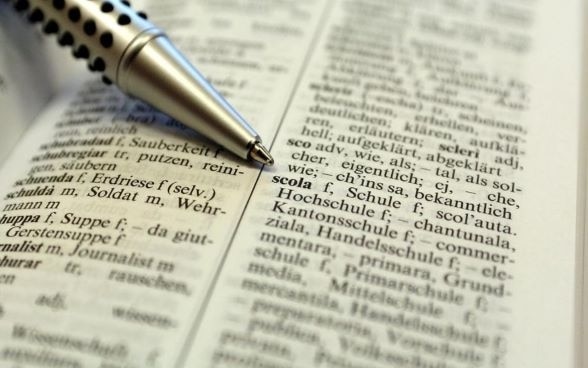 Открытая страница ретороманского словаря, на которой ручка указывает на слово «scola», означающее на ретороманском «школа». 