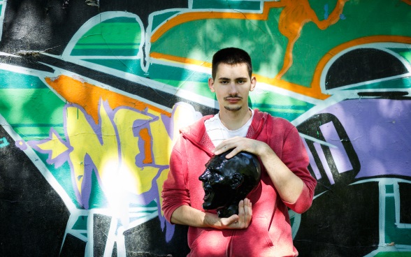 A young man standing in front of a graffiti-covered wall, holding a sculpture of a head in his hands.