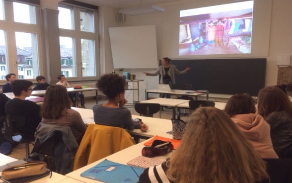 Eine Frau steht vor einer Schulklasse und spricht zu den Schülerinnen und Schülern, an die Wand sind Fotos projiziert. 