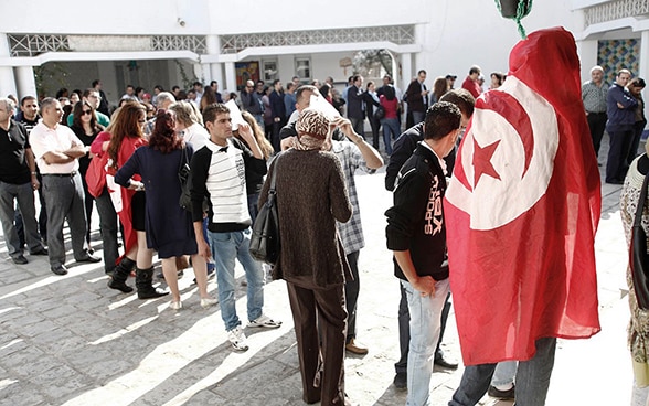  Une longue file d'attente devant un bureau de vote en Tunisie. Une personne au premier plan est enveloppée dans un drapeau national tunisien.