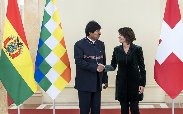 Evo Morales Ayma und Doris Leuthard schütteln sich vor den Landesflaggen die Hand.