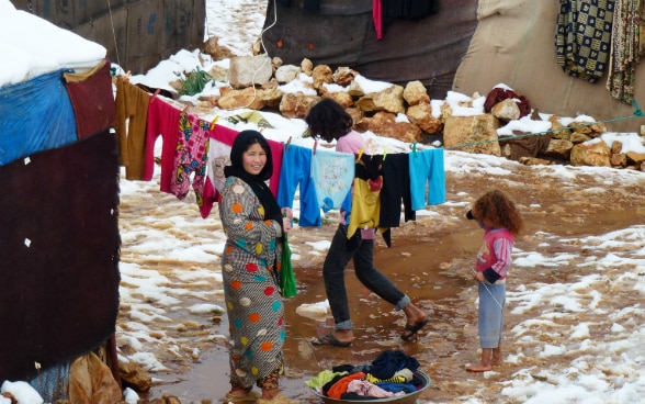 Una donna in un campo profughi stende i panni tra due tende. Accanto a lei ci sono due bambini. Per terra vi è uno strato di neve.
