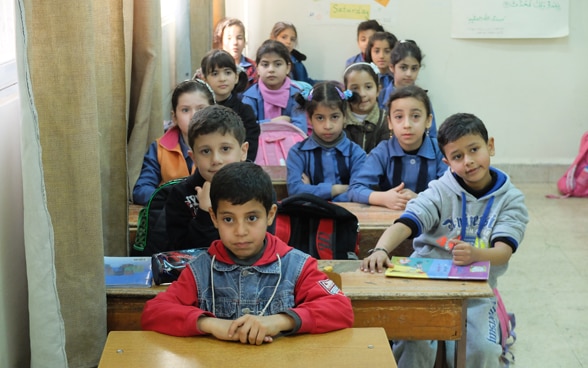 A group of girls and boys sit tightly packed at desks in a classroom.