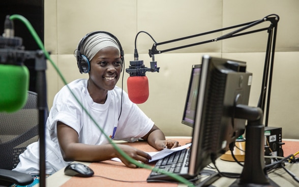 A female African radio presenter makes an entertaining radio show. She uses a red microphone and a computer.