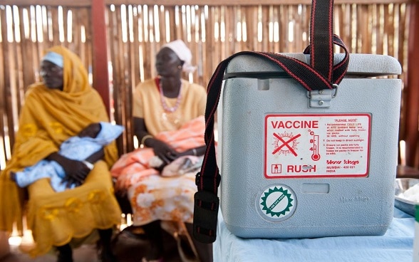 Assises sur des chaises, deux femmes soudanaises attendent leur tour avec leur nourrisson dans les bras. Au premier plan, on aperçoit un conteneur frigorifique dans lequel sont stockés des vaccins.