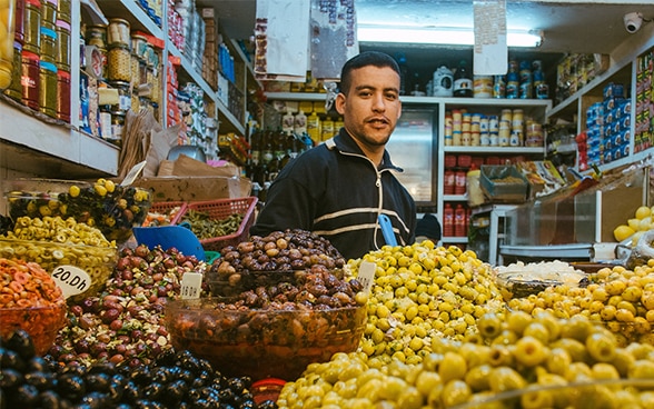 The picture shows a man in a small shop in front of a large display of olives.