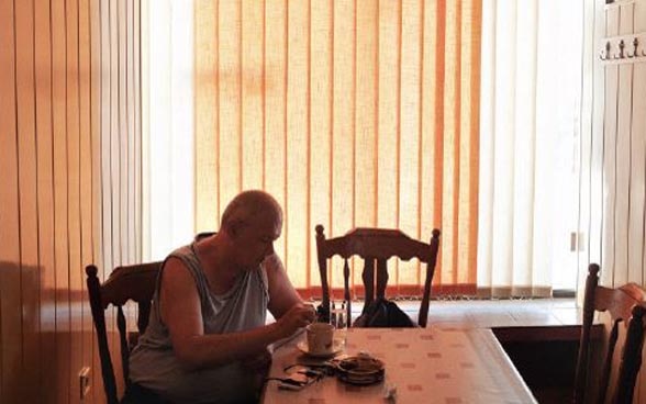 An elderly man sitting alone at a table in a dimly-lit room.