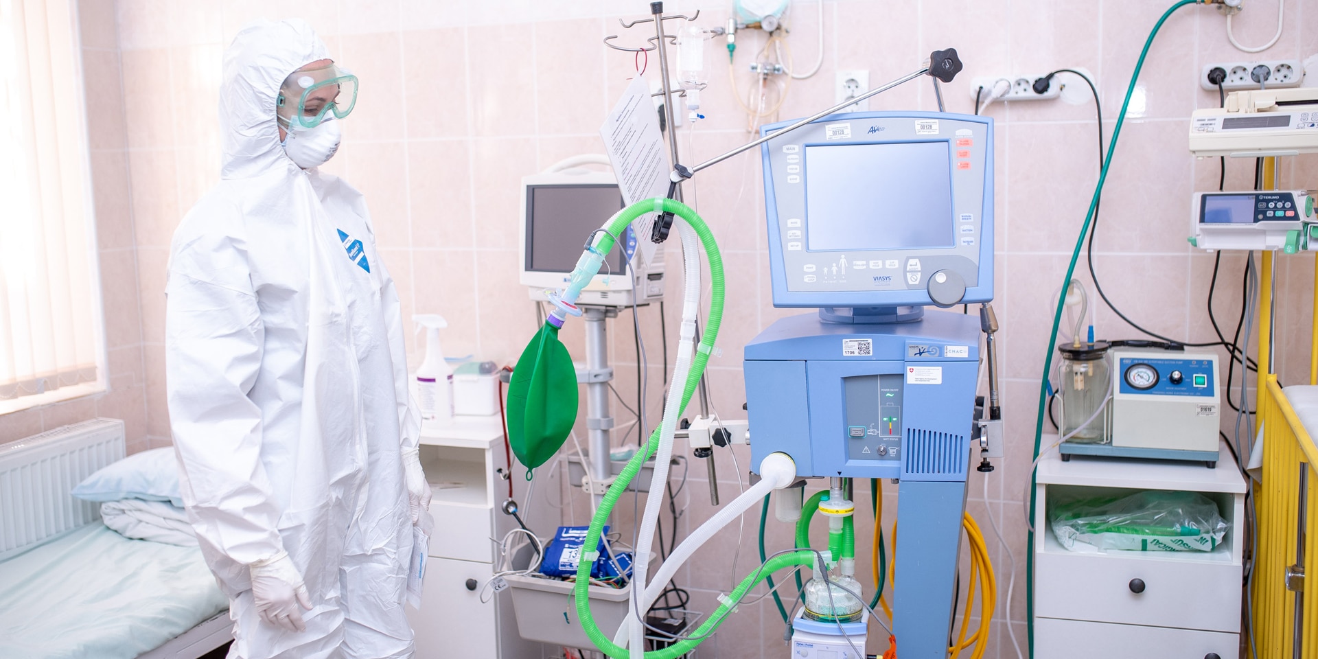 Eine Frau, die einen Schutzanzug, eine Brille und eine Maske trägt, steht in einem Krankenzimmer neben medizinischen Apparaten. 