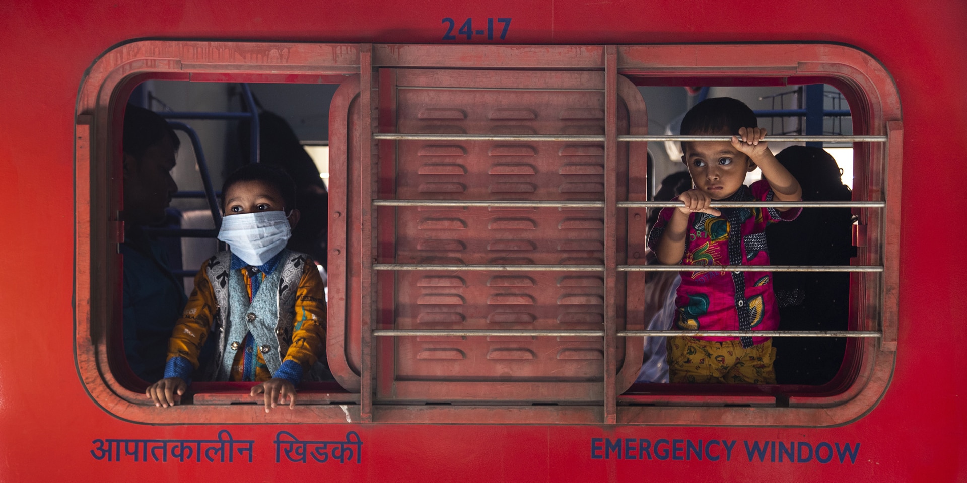 Deux enfants, l’un avec masque, l’autre sans, derrière la vitre coulissante d’un guichet.