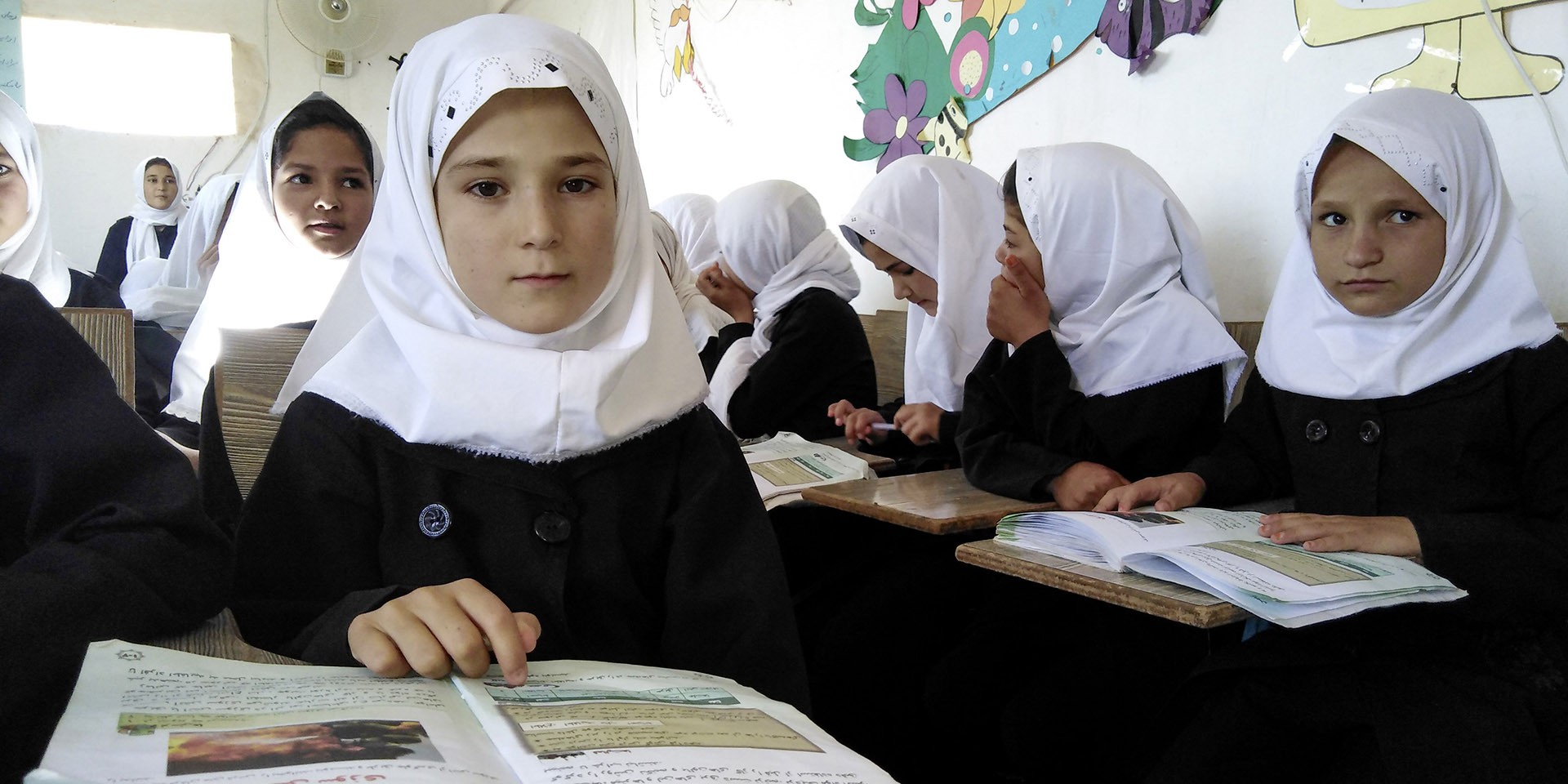 Einige Mädchen mit Kopftüchern im Vordergrund. Sie befinden sich in einem Klassenzimmer und lesen Schulbücher. 