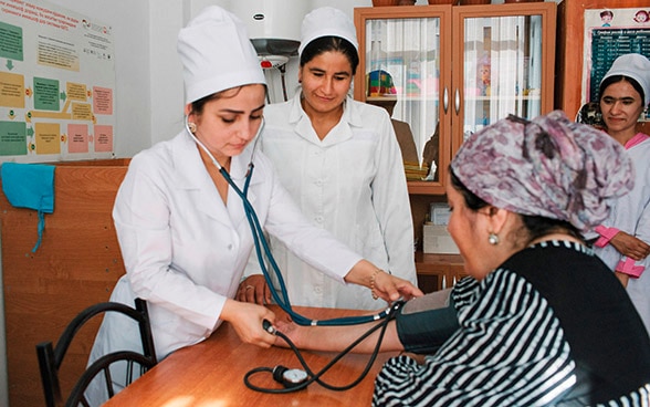 Zwei tadschikische Ärztinnen messen den Blutdruck einer Patientin, der auf einem Stuhl sitzt.