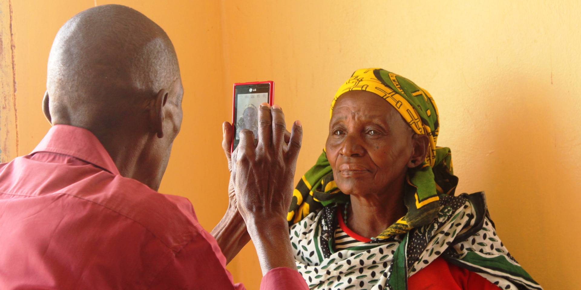 Un uomo scatta una foto ritratto con il suo cellulare a una donna. La donna è in piedi davanti a una parete arancione. 