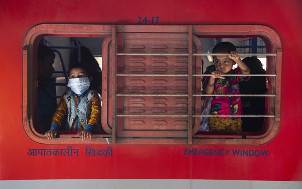 Zwei Kinder schauen aus dem Schiebefenster eines Schalters – eines mit, eines ohne Maske.