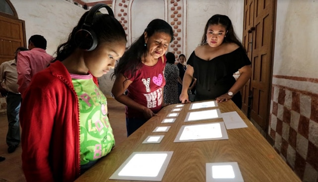 A little girl with headphones is watching screens on a table. Next to her are two women. 