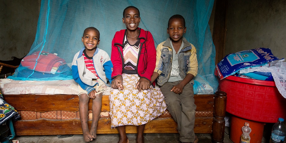 A woman is sitting on a bed with her two children. The bed is protected by a mosquito net.