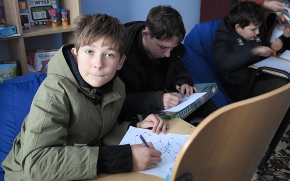 Un garçon assis dans une pièce écrit sur une feuille de papier posée sur une chaise devant lui. Autour de lui, d’autres enfants apprennent leurs leçons dans une position similaire. 