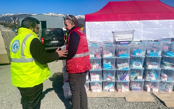 Une femme membre du Corps Suisse d’aide humanitaire vêtue gilet rouge ainsi qu’un homme avec un gilet jaune se tiennent debout devant des caisses contenant des kits d’hygiène.