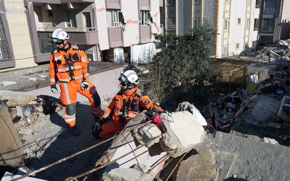 Two men in orange clothes stand on the rubble of a collapsed house.