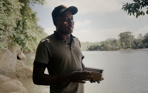 A man is holding a packet of chocolate, standing at a river bank.