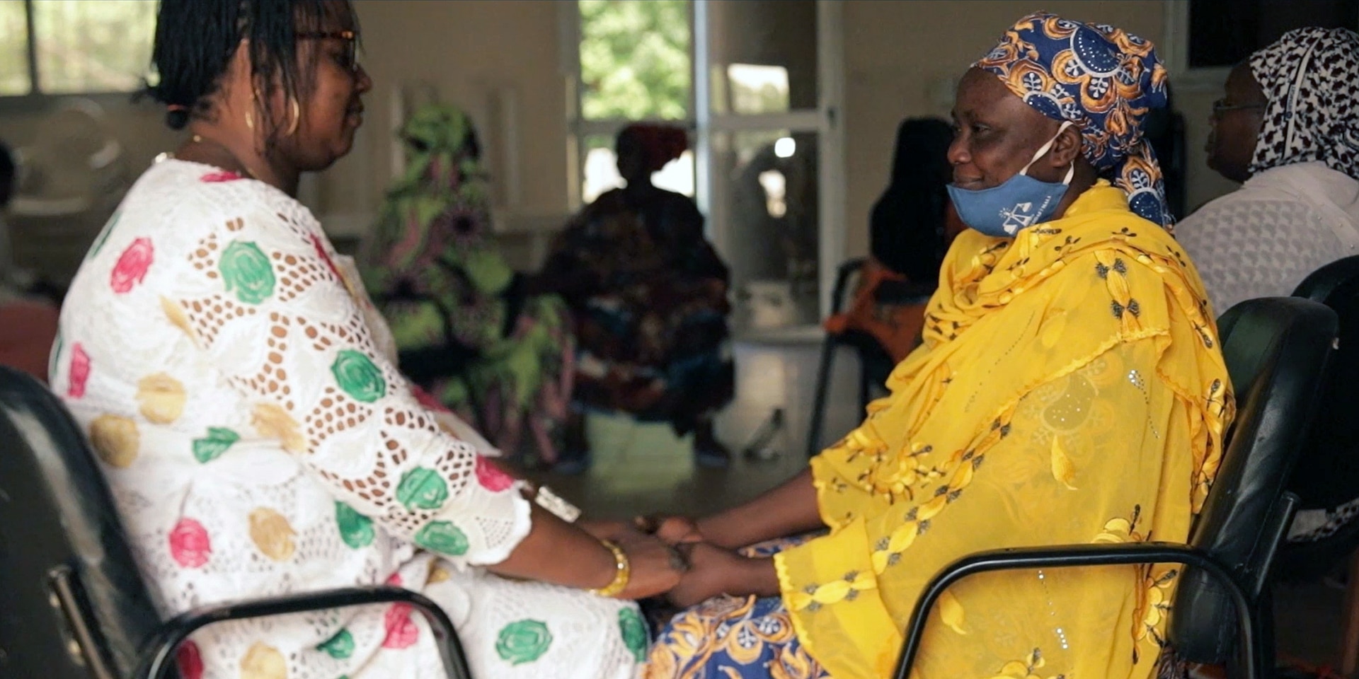 Two women are sitting across from each other, looking at each other and holding hands