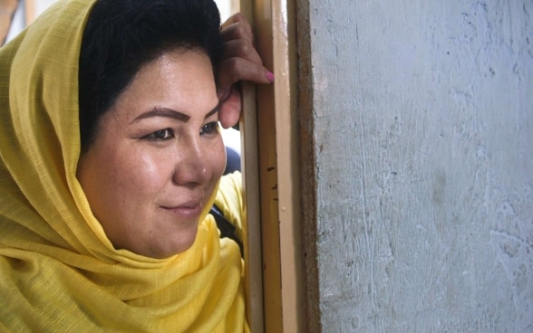 A smiling Afghan woman wearing a traditional yellow headscarf leans on a door frame.