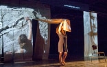 An artist, dressed in a brown dress, stands on a stage. She wears a wide band that she holds over her head and connects her to an image projected on her back.