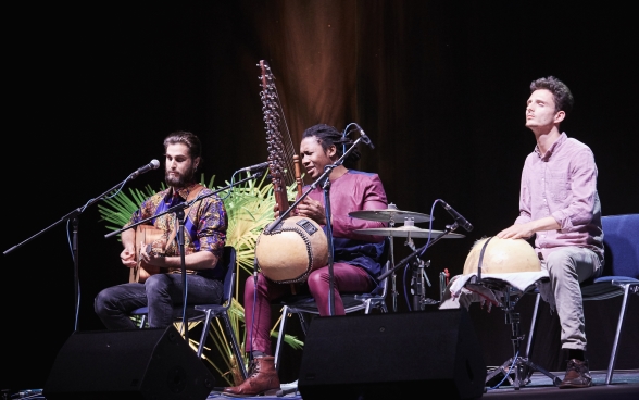 Drei Musiker sitzen mit ihren Instrumenten auf der Bühne vor ihren Mikrophonen. Der Musiker in der Mitte singt.