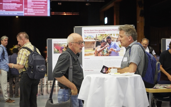 Deux participants sont debout à une table de la Festhalle de Bernexpo. Derrière eux, on voit l’exposition des organisations partenaires.