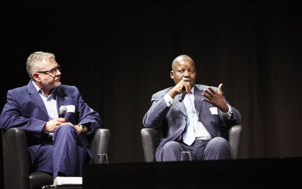 Mansour N’Diaye est assis sur la scène et parle dans son microphone. Dominique Guenat est assis à côté et écoute attentivement.