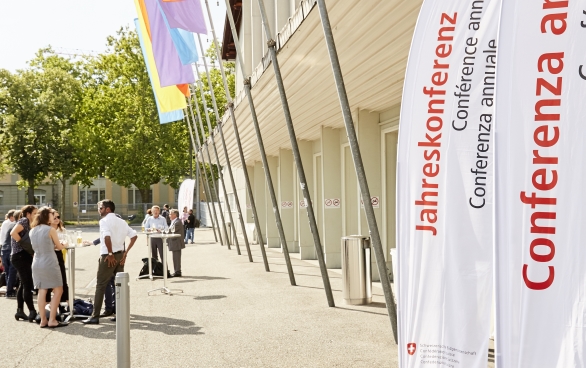 The Bernexpo Festhalle from outside. Colourful flags are hanging in front of the doors and there are white posters bearing the word 'Jahreskonferenz' in all the official languages and English. 