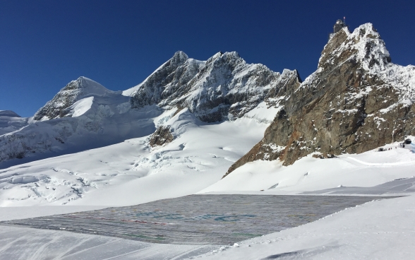 La più grande cartolina del mondo esposta sul ghiacciaio; sullo sfondo, alcune vette. 