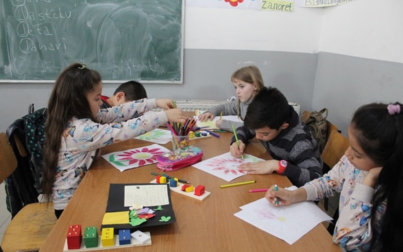 Des filles et des garçons sont en classe, assis autour d’une table devant le tableau. Ils font chacun un dessin coloré.