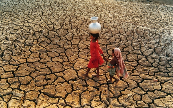 Two children with a water jug walk through a dried out riverbed.
