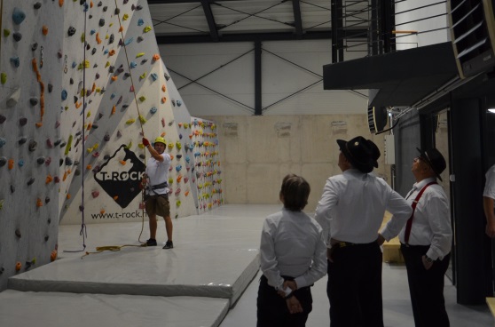 Tre musicisti svizzeri in costume tradizionale osservano due uomini scalare una parete in una palestra di arrampicata.