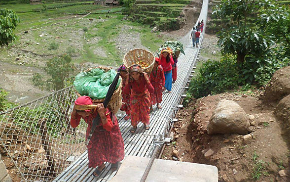 Women carrying baskets walk on a trail bridge.