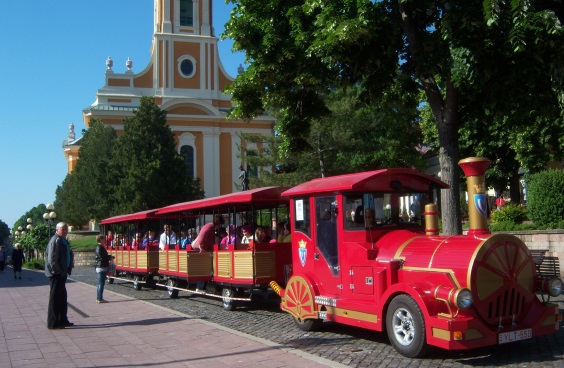 Ein kleiner, roter Tourismuszug mit einer Lokomotive und drei Waggons tuckert über eine Strasse aus Kopfsteinpflaster.