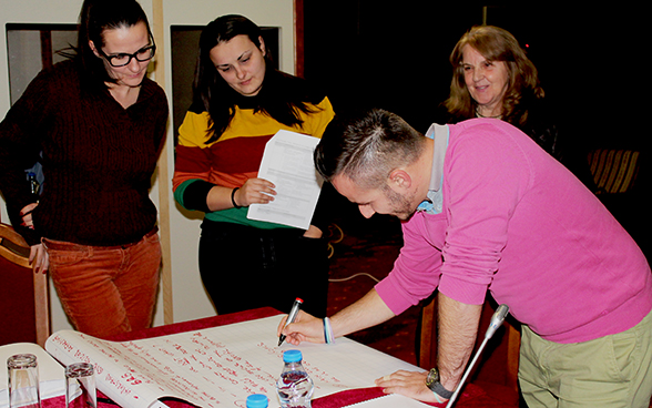 Three women look on as a young man writes something on a piece of paper.