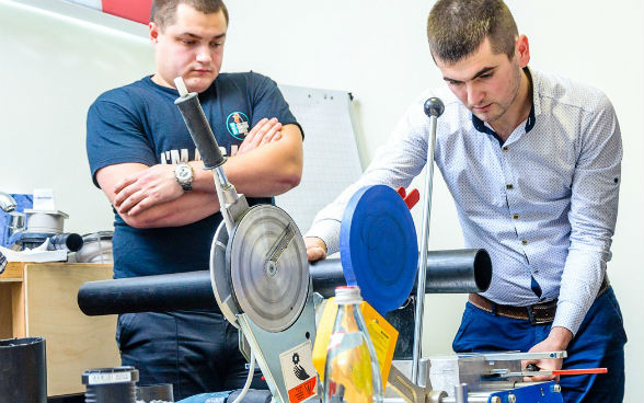 Two plumbers are cutting a pipe with a machine in a workshop.