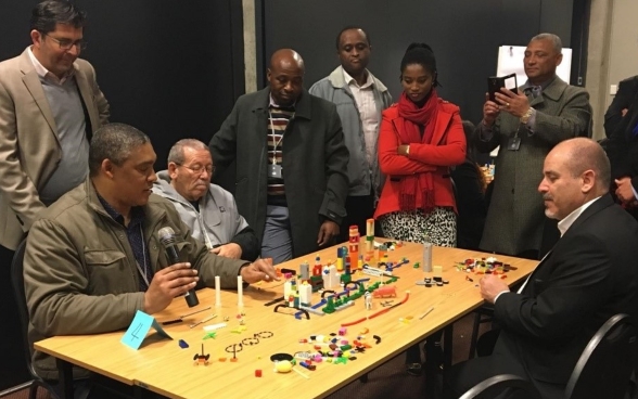 Un groupe de personnes est attablé devant la maquette d’une ville constituée de pièces colorées. Un homme, microphone à la main, désigne un élément de la maquette.