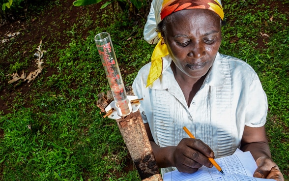 A young Kenyan farmer named Mercy Wambui stands on the grass. She measures rainwater and records the data on her list.
