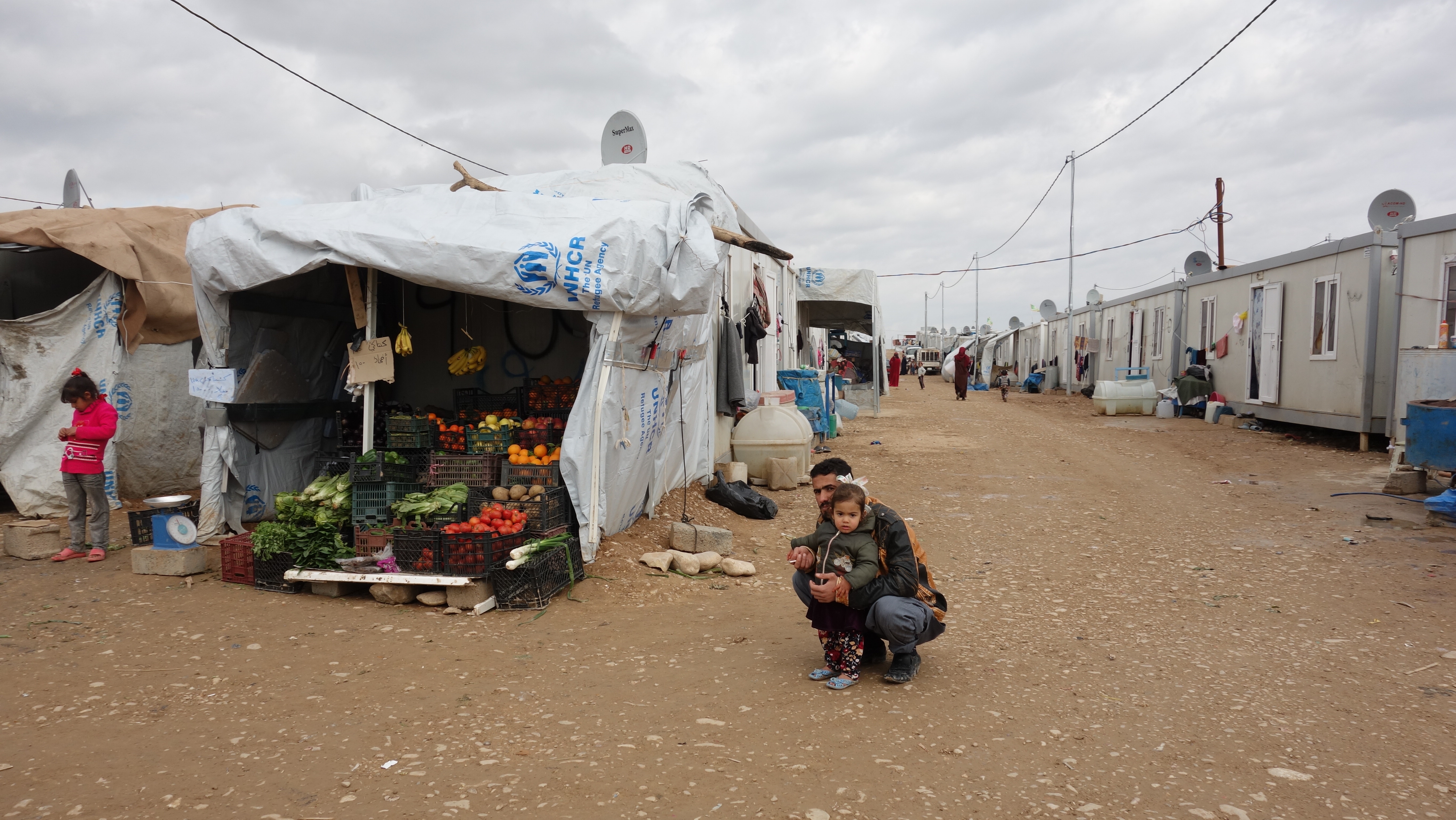 Ein Mädchen und sein Vater neben einem UNHCR-Zelt mit Früchten und Gemüse. 