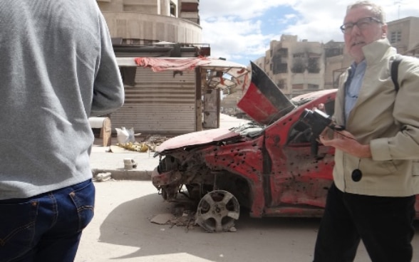 Henri Stalder stands in front of a wrecked car riddled with bullet holes.