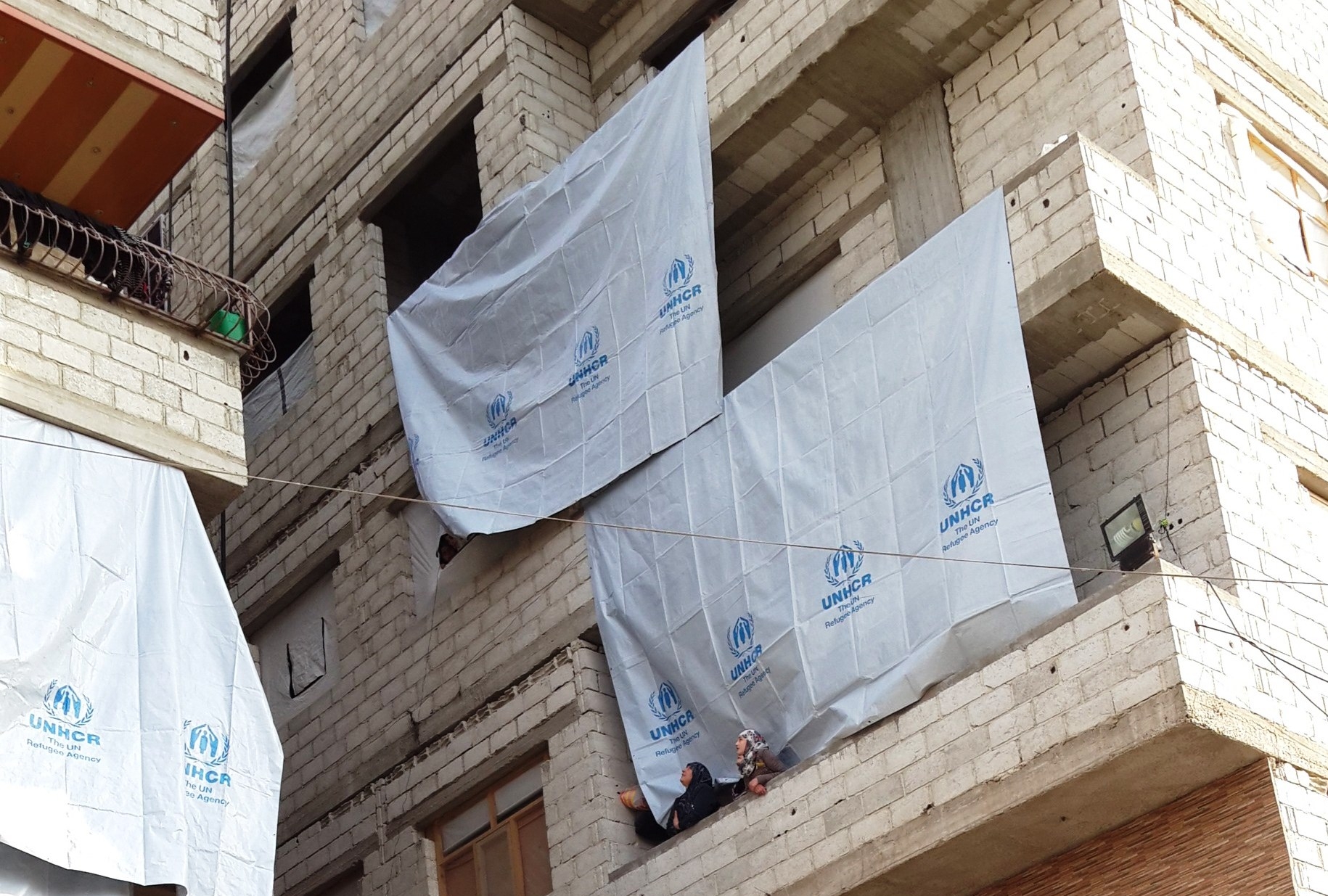 Un edificio con banderas del ACNUR y dos mujeres con velo en un balcón. 