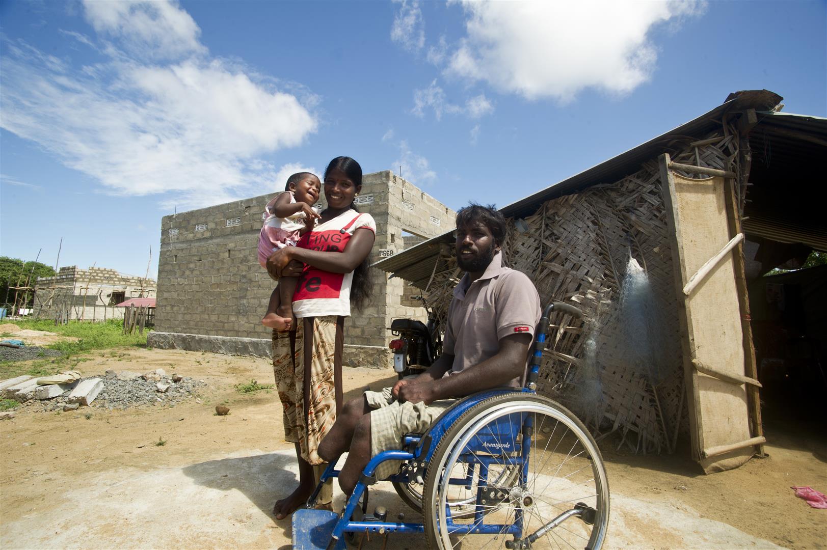 Un hombre en silla de ruedas, su esposa y su hijita, delante de su casa.