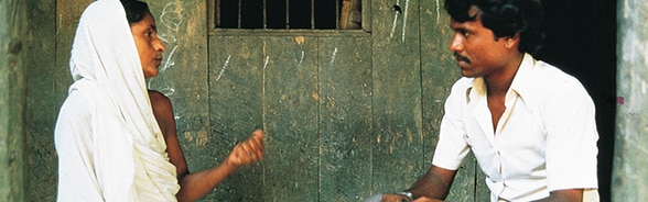 A woman and a man talk outside a hut.