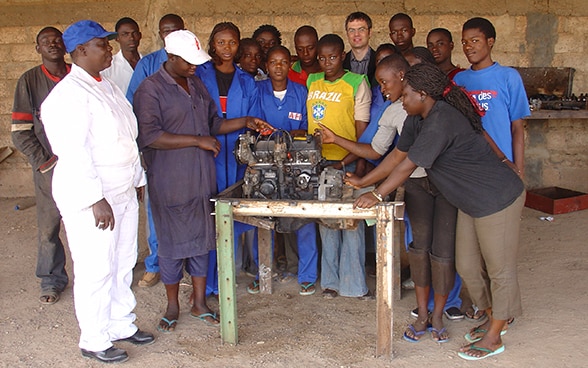 Women and men in Burkina Faso stand around an engine on a table.