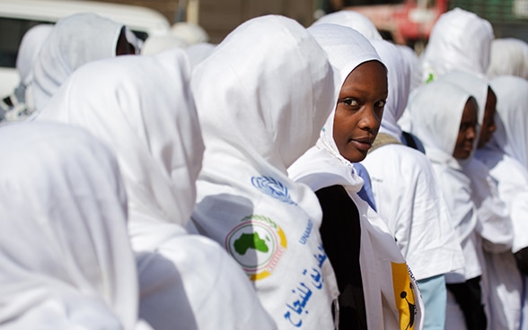 Girls wearing white head scarves stand in a row; one girl looks back.