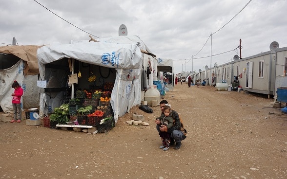 A father and daughter beside a fruit and vegetable stall in a refugee camp.