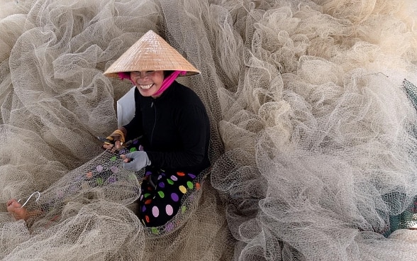A smiling young woman sits among the fishing nets with a tool in one hand and a piece of net that she is repairing in the other. 